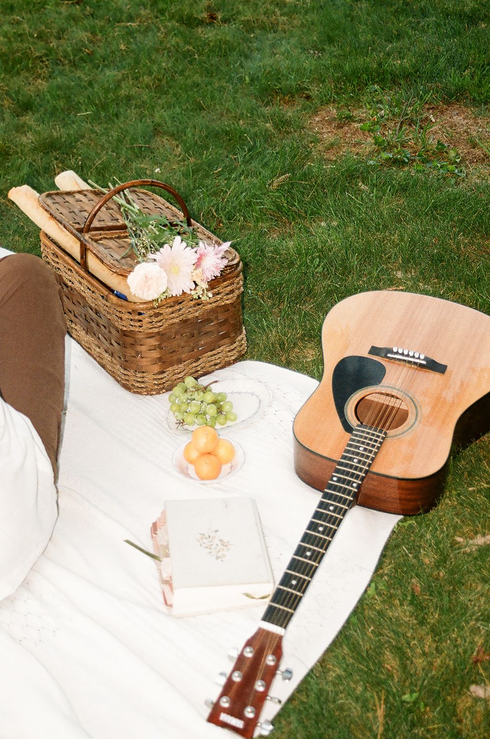 Romantic outdoor picnic with wicker basket, fresh flowers, acoustic guitar, fruit, and journal on white blanket in green grass.