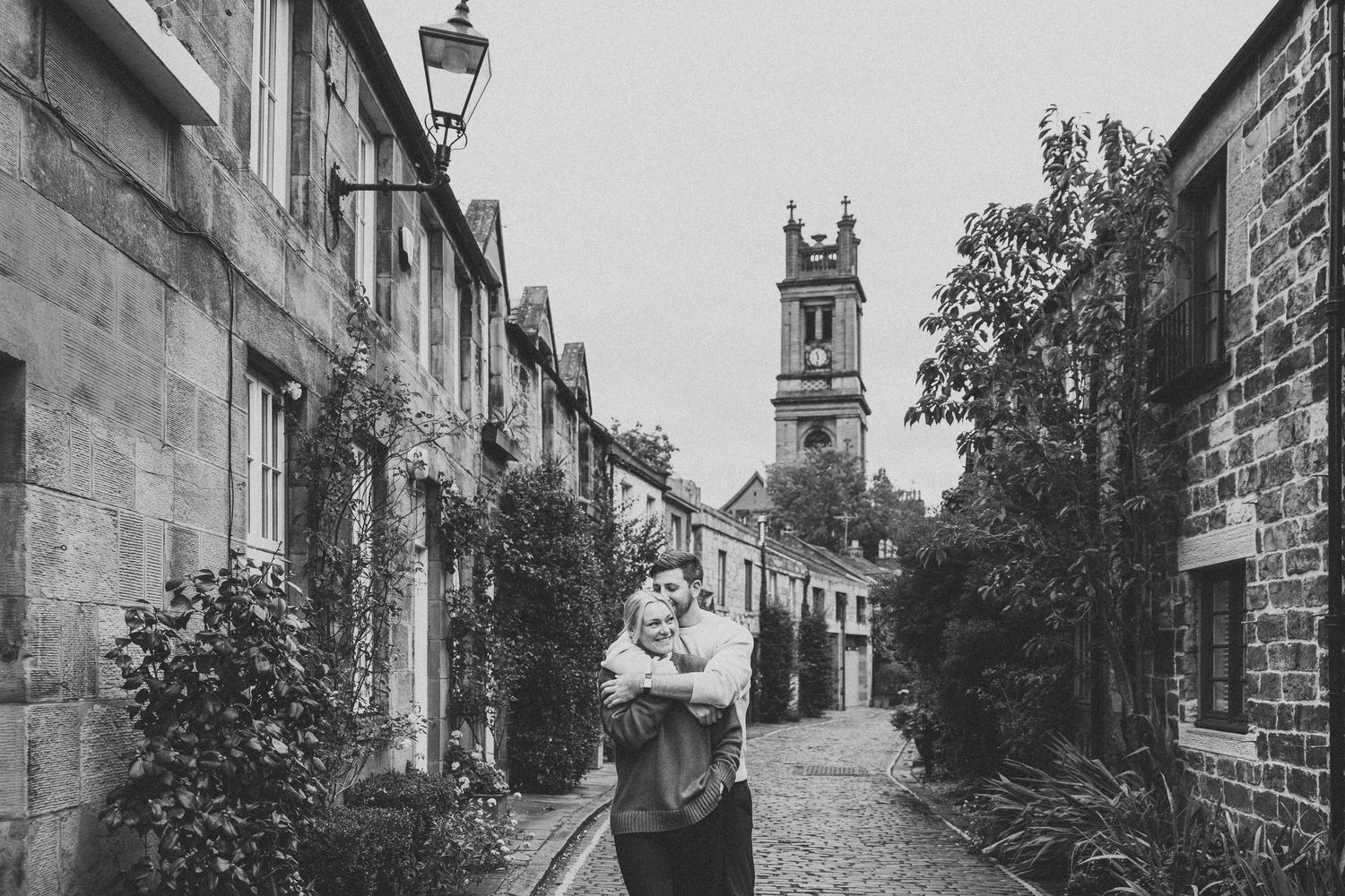 Couple embracing on a charming cobblestone alley with stone buildings and a church tower in black and white.