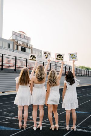Four friends in white dresses hold up university signs with their backs turned on an athletic track field.