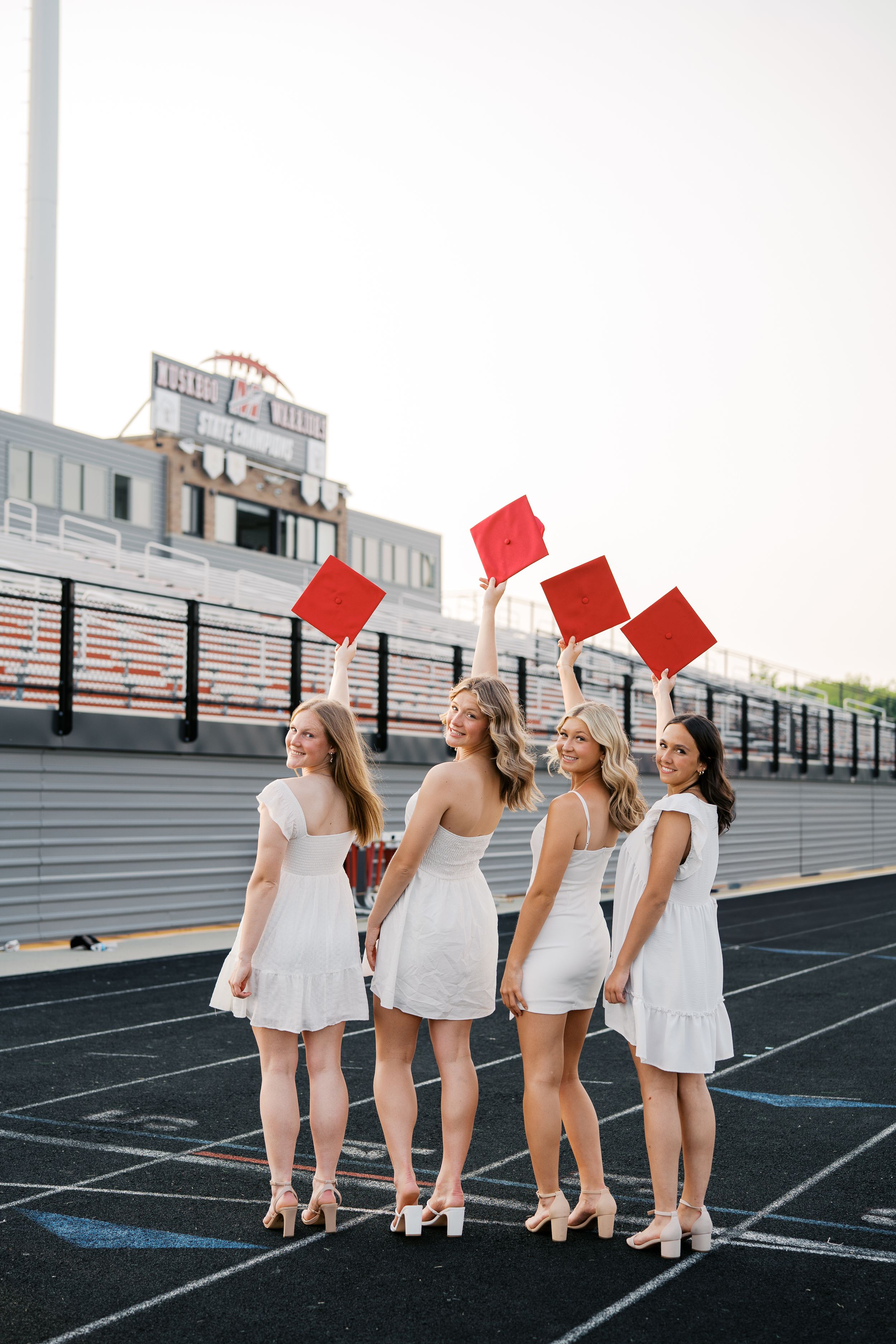 Four graduates in white dresses hold up red diplomas while posing on track field near stadium bleachers.