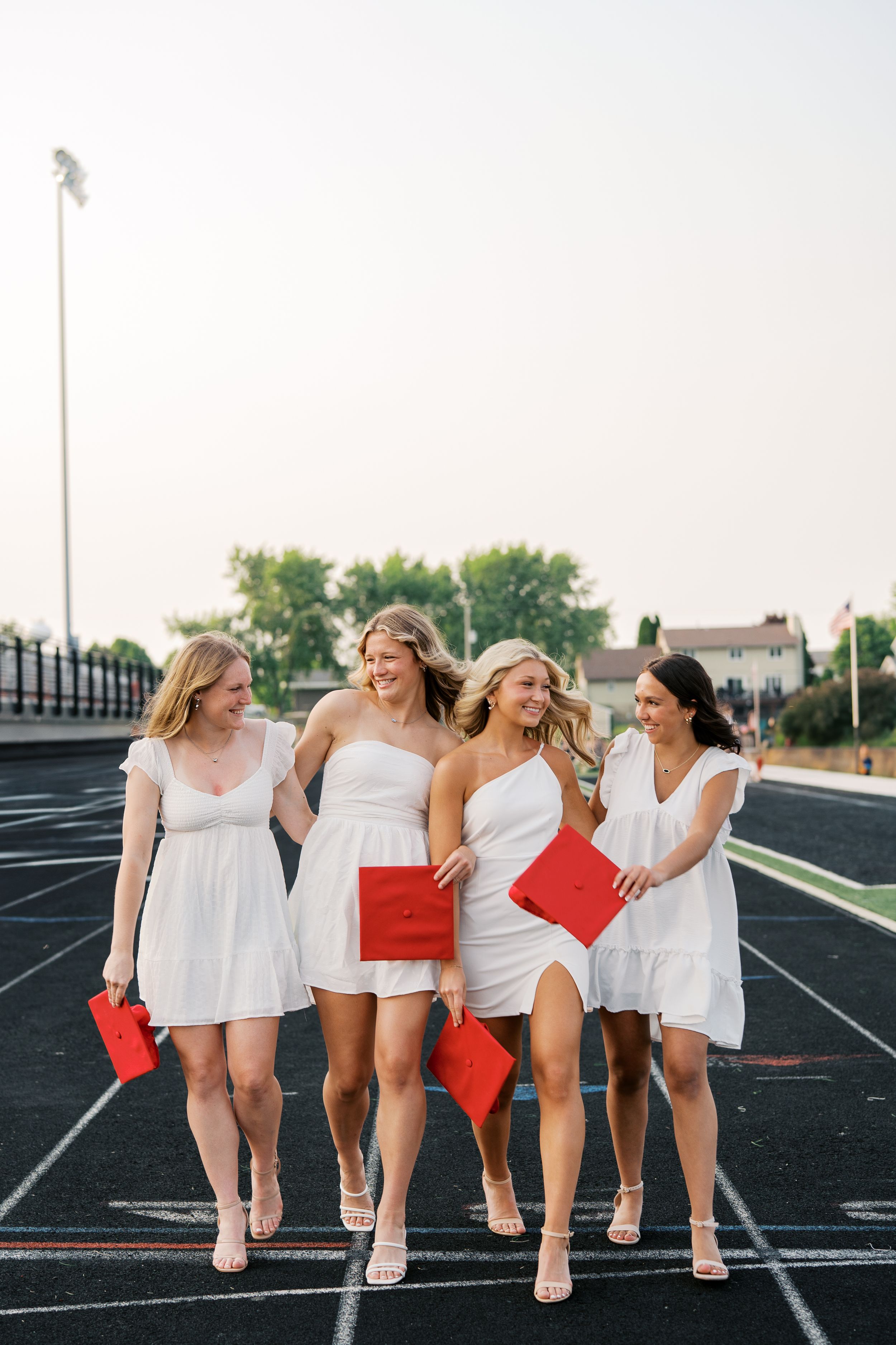 Friends in white dresses sitting together on track field lines while laughing and embracing.