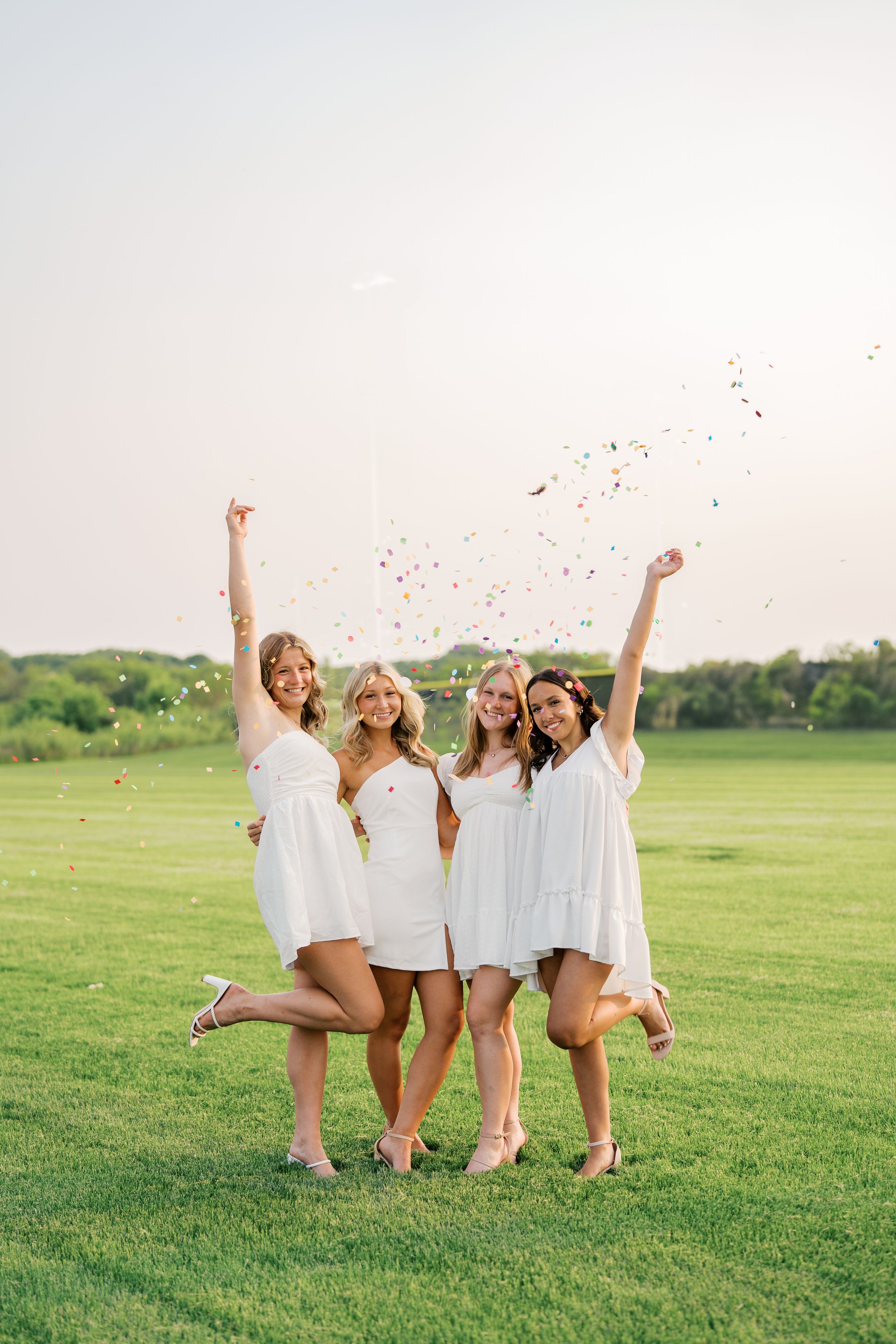 Group celebrates on green field in white dresses throwing confetti in the air.