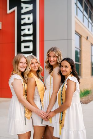 Four graduates in white dresses pose together with honor cords against red and white building exterior.
