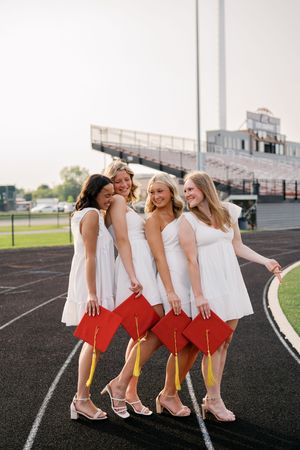 Group of friends in white dresses and red skirts pose playfully on a track field showing off their matching outfits.