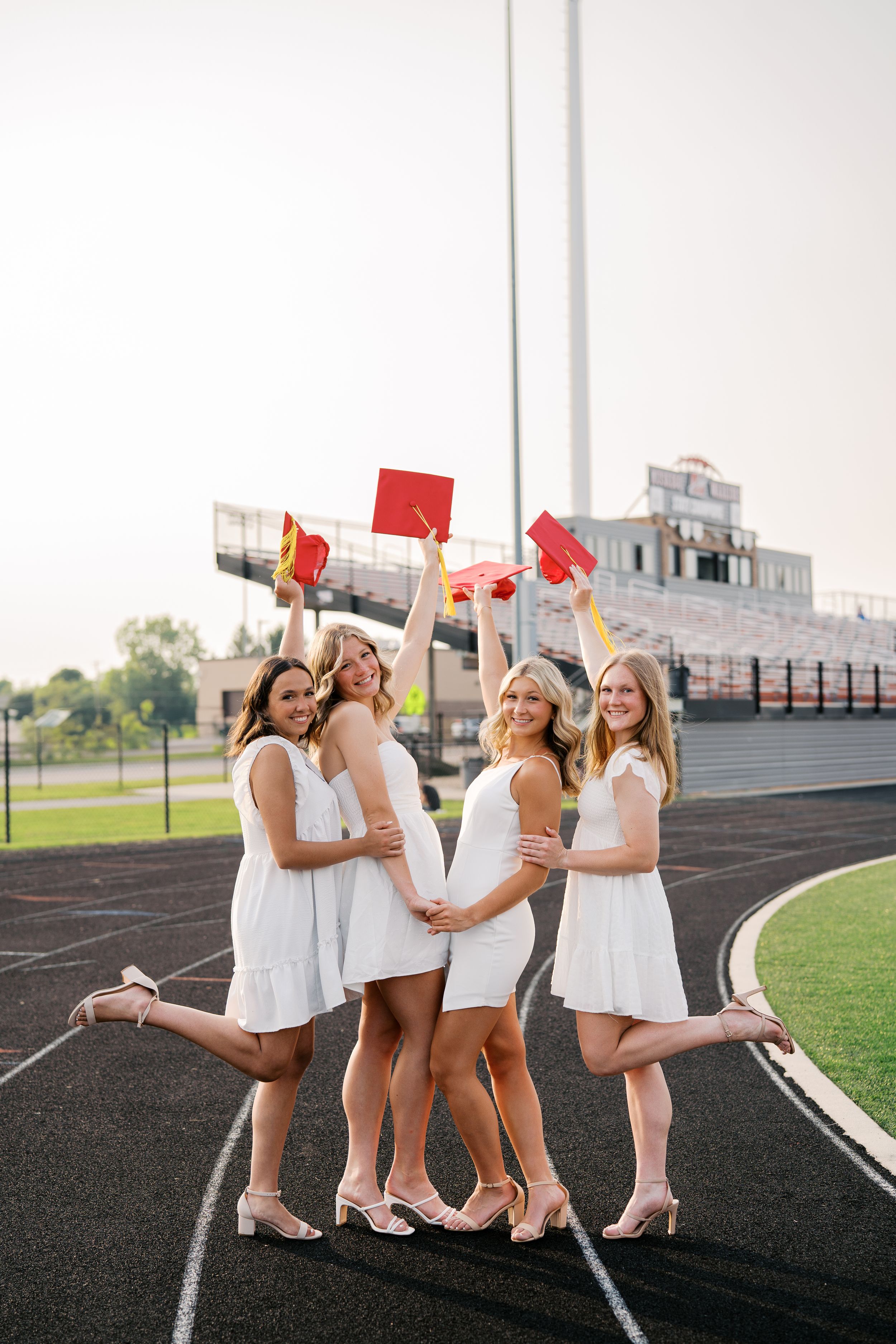 Four graduates in white dresses celebrate on a track field while holding red diplomas and wearing matching heels.