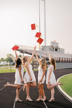 Group in white dresses holding red folders while posing playfully on a track field with stadium bleachers.