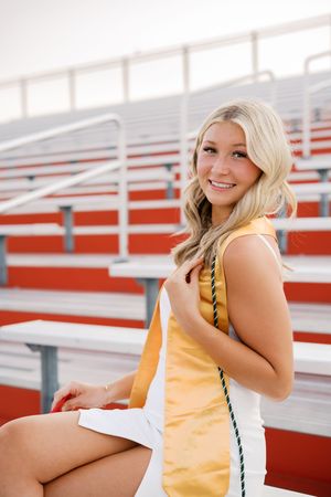 Person in yellow cheerleader uniform sitting on red stadium bleachers during a sunny day.