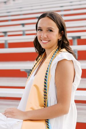 Graduate in white dress and honor cords poses on stadium bleachers with red and white stripes.