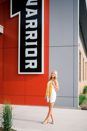 A person in a white outfit and yellow top poses against a modern building exterior with red wall and Warrior signage.