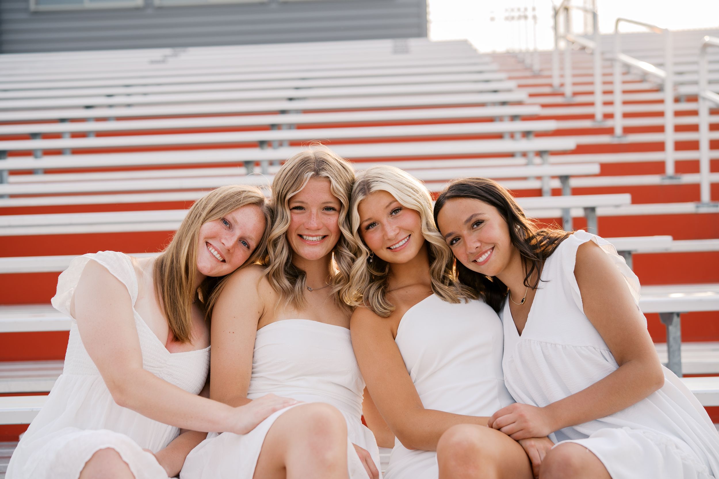 A group of cheerleaders in white uniforms pose together on red stadium bleachers for a team photo.
