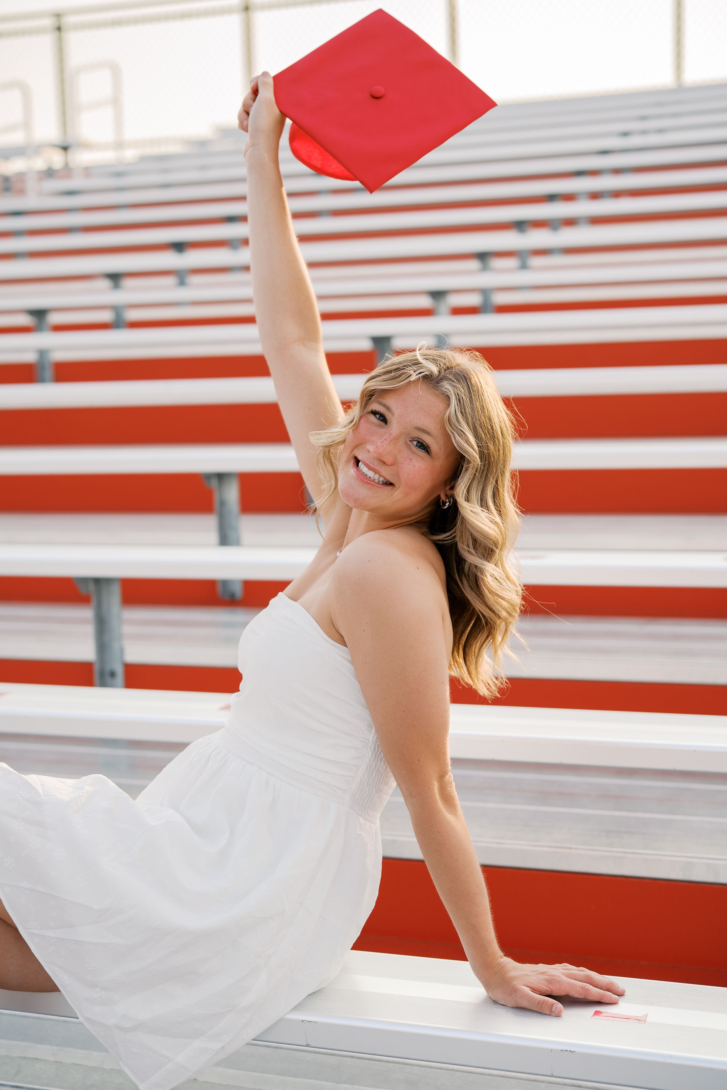 Person in white dress posing with red graduation cap on stadium bleachers.
