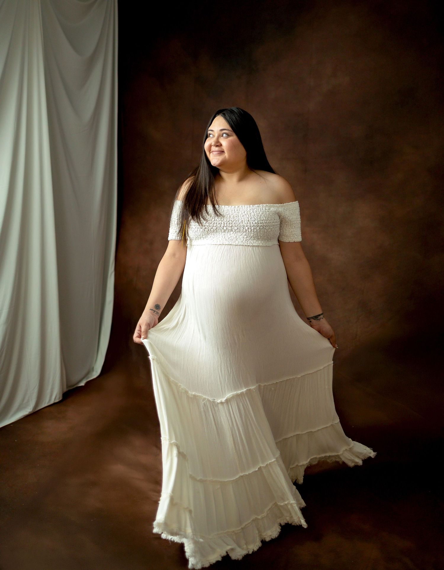 Pregnant woman in sheer white ruffled mini dress with bell sleeves, posing gracefully against gray background.