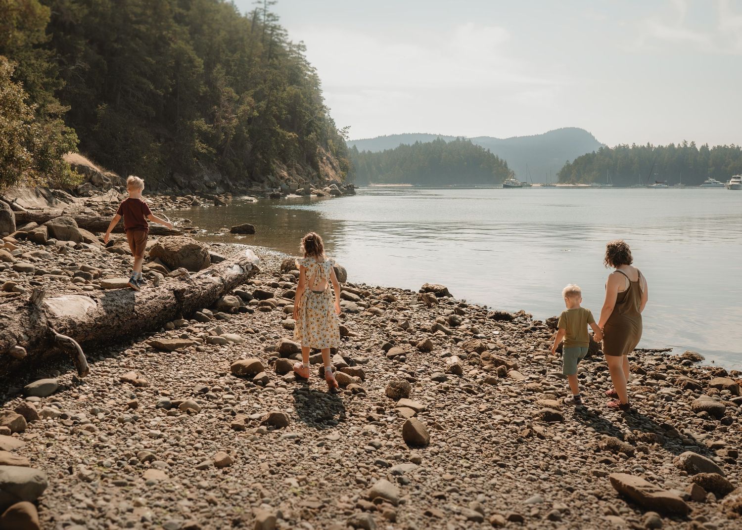 Family with children exploring a rocky shoreline beside a calm river surrounded by forested mountains.