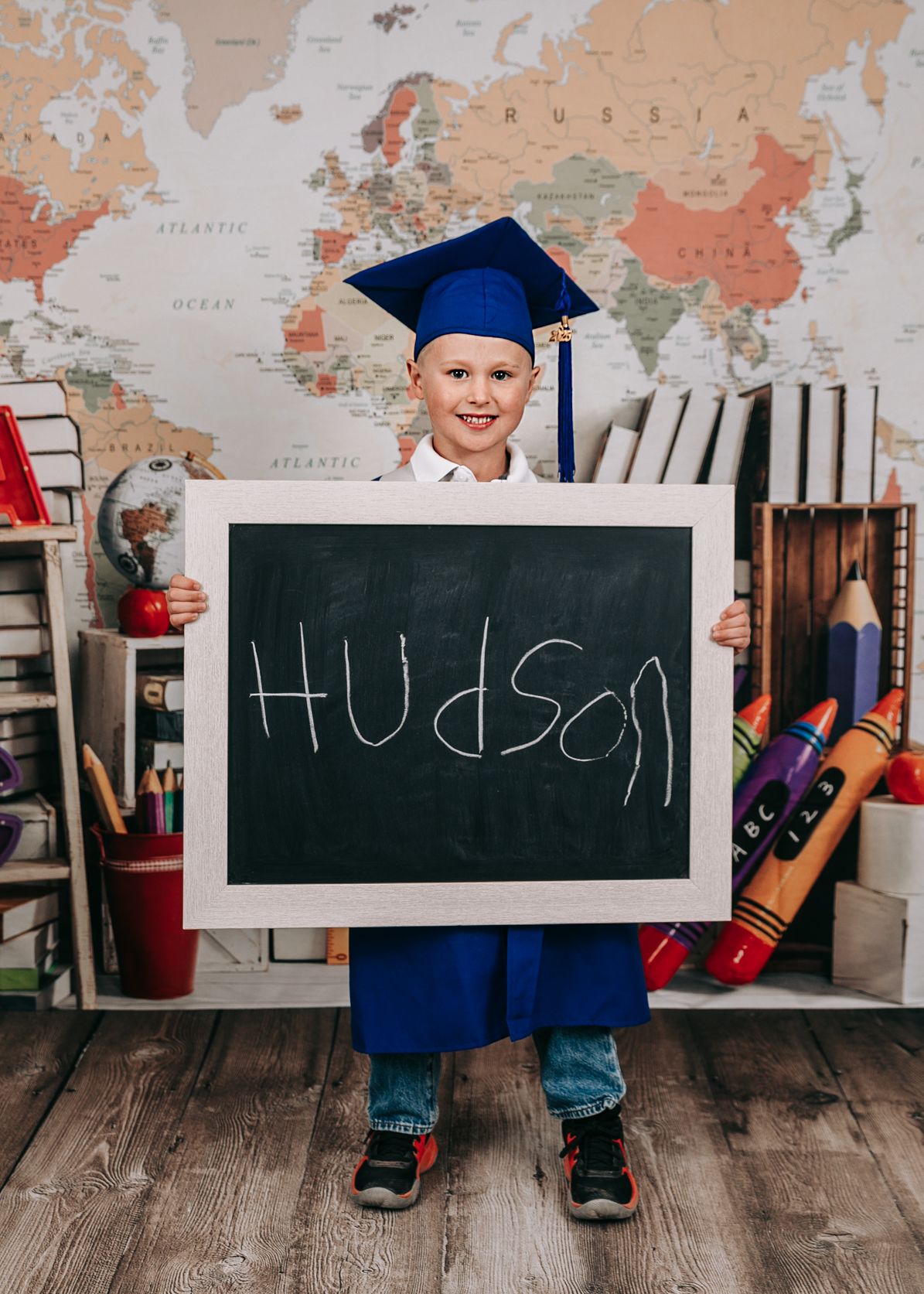 A young boy in a blue graduation cap and gown holds a chalkboard sign spelling his name Hudson.