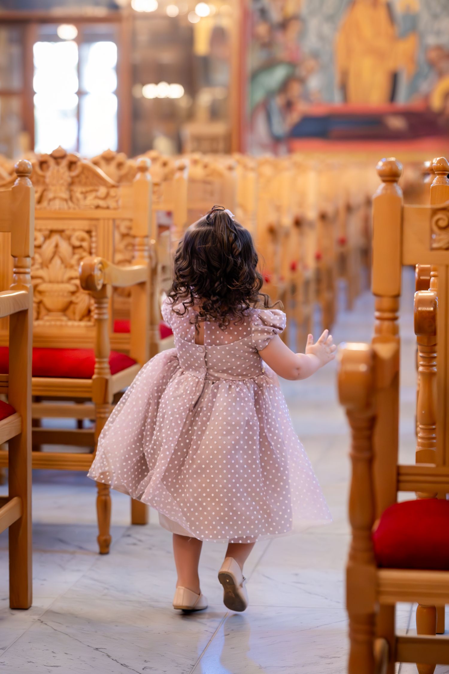 A young girl in a polka dot dress walks down the aisle of an ornate wooden church interior.