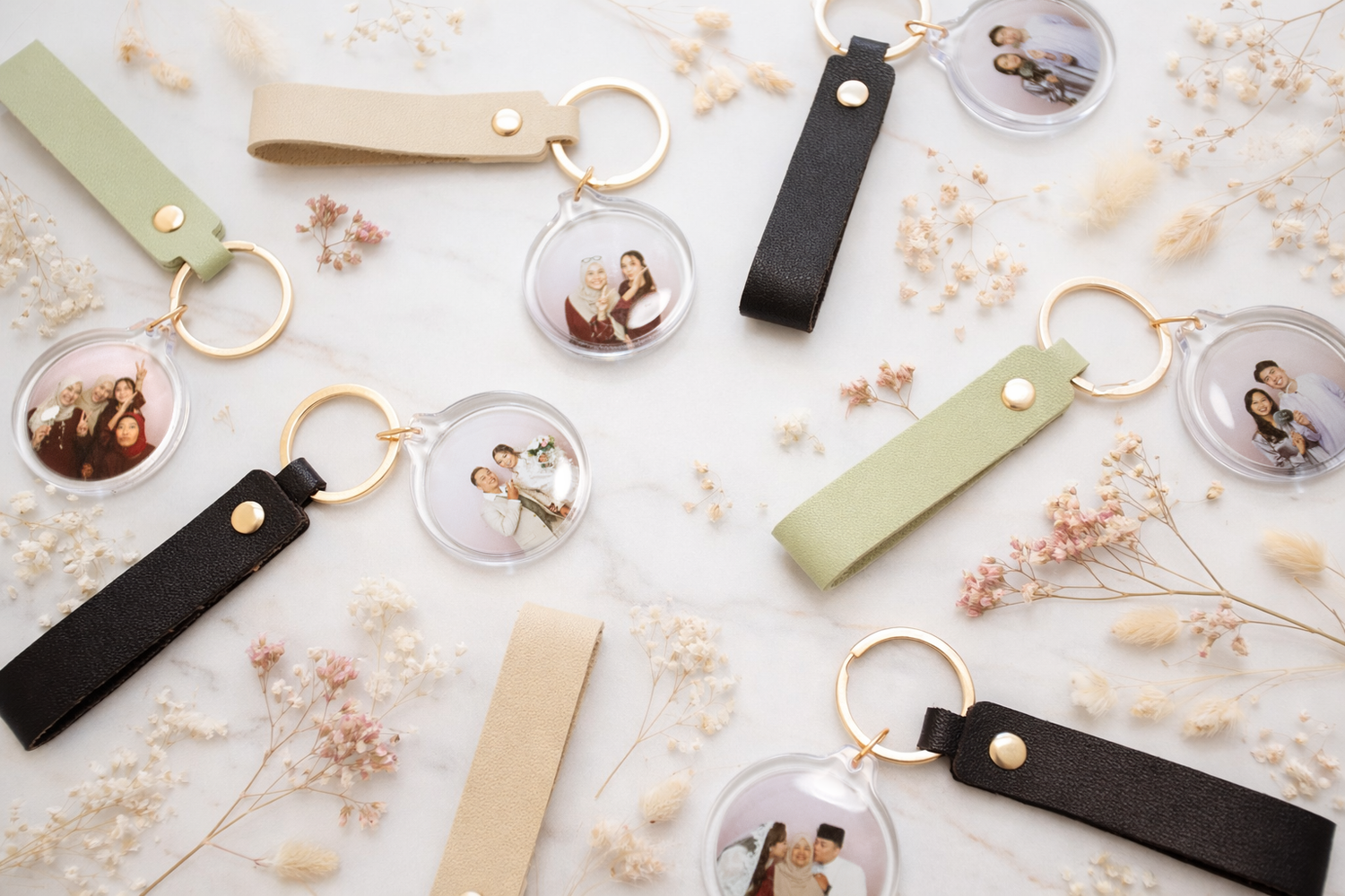 Keychains with round photo inserts and leather straps are arranged on a white surface decorated with small dried flowers.