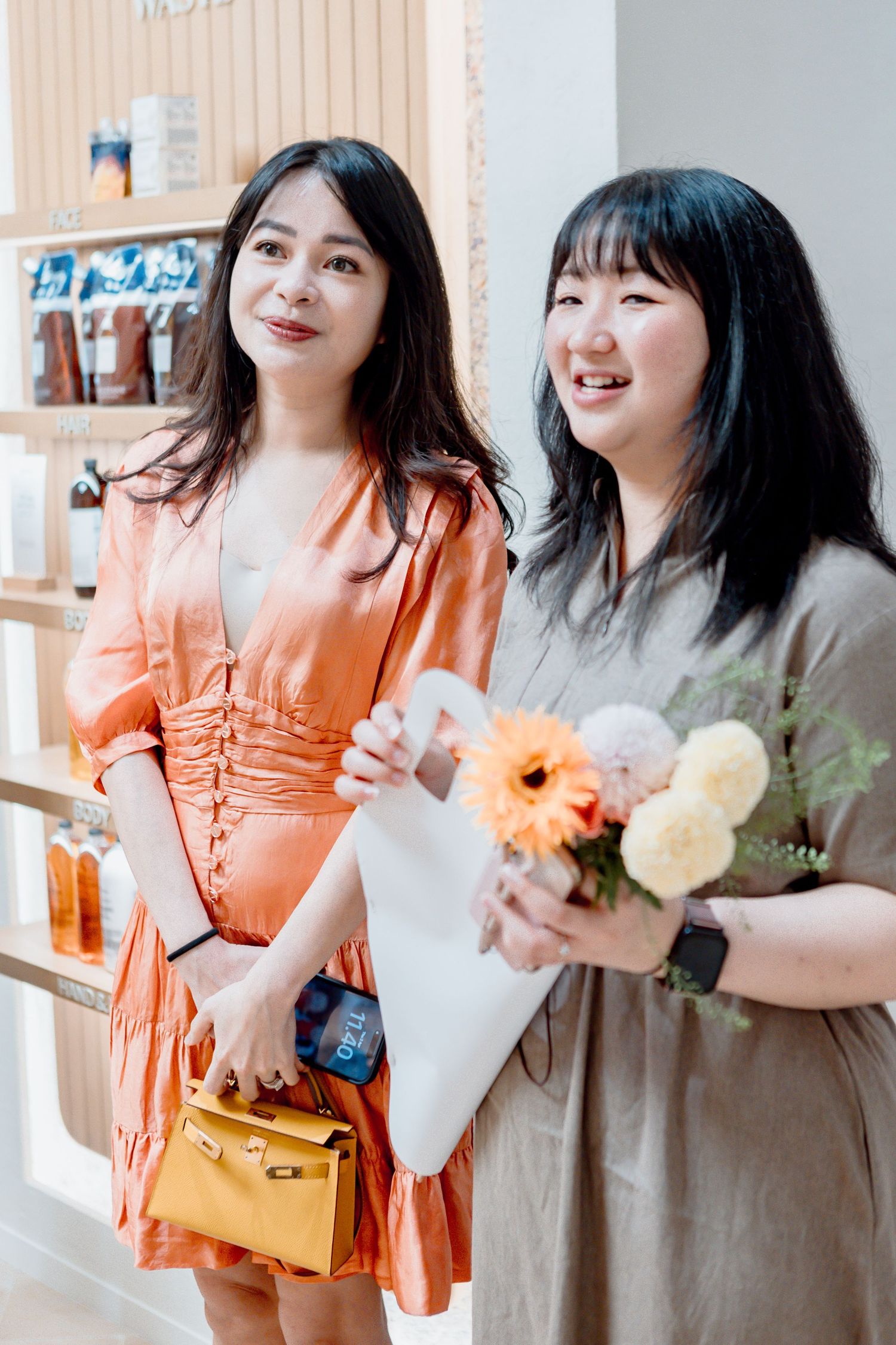 Two Asian women smiling in a shop, one in orange dress with yellow bag, other in gray dress holding flowers.