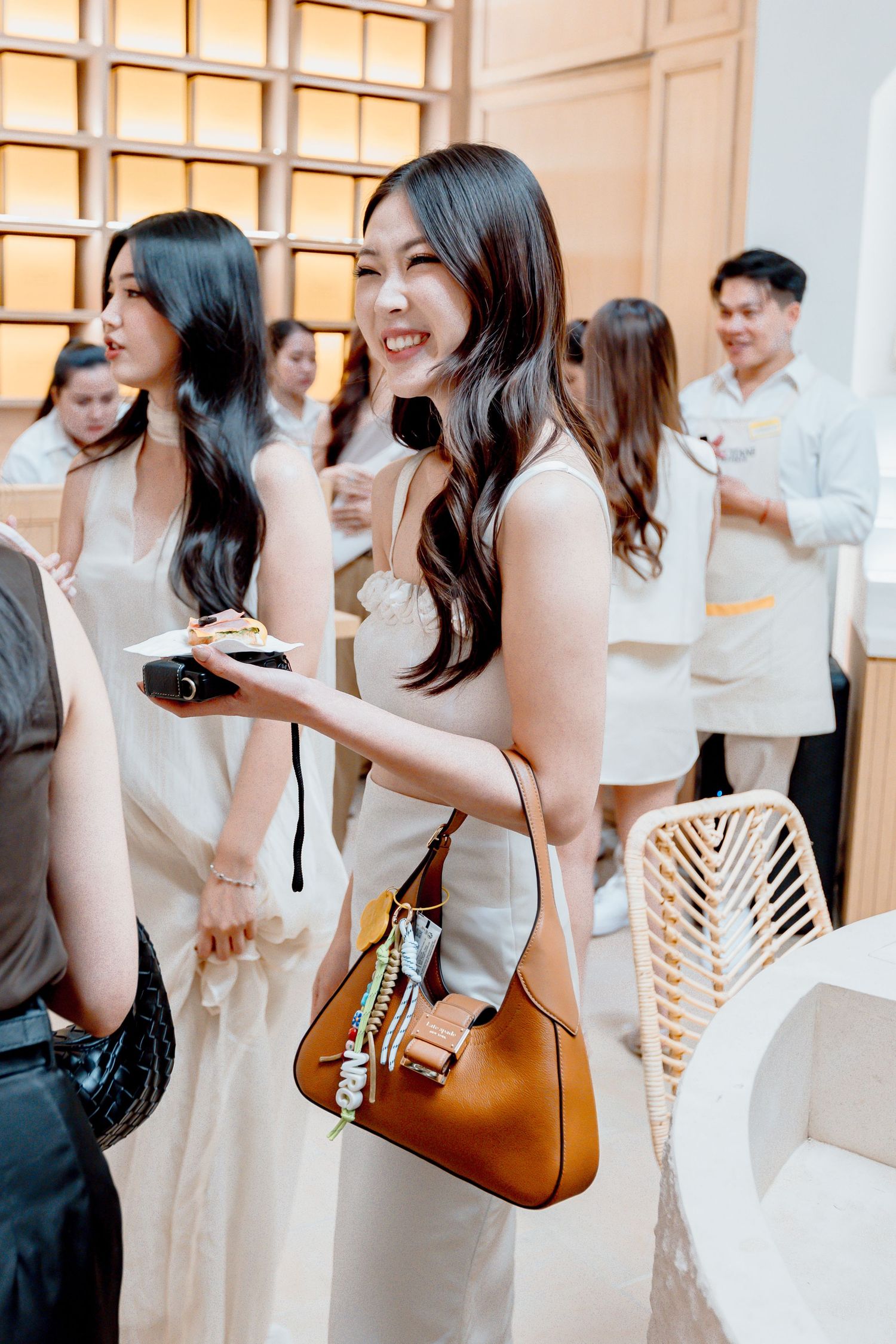 Smiling Asian woman in white dress with brown handbag at elegant social gathering event.