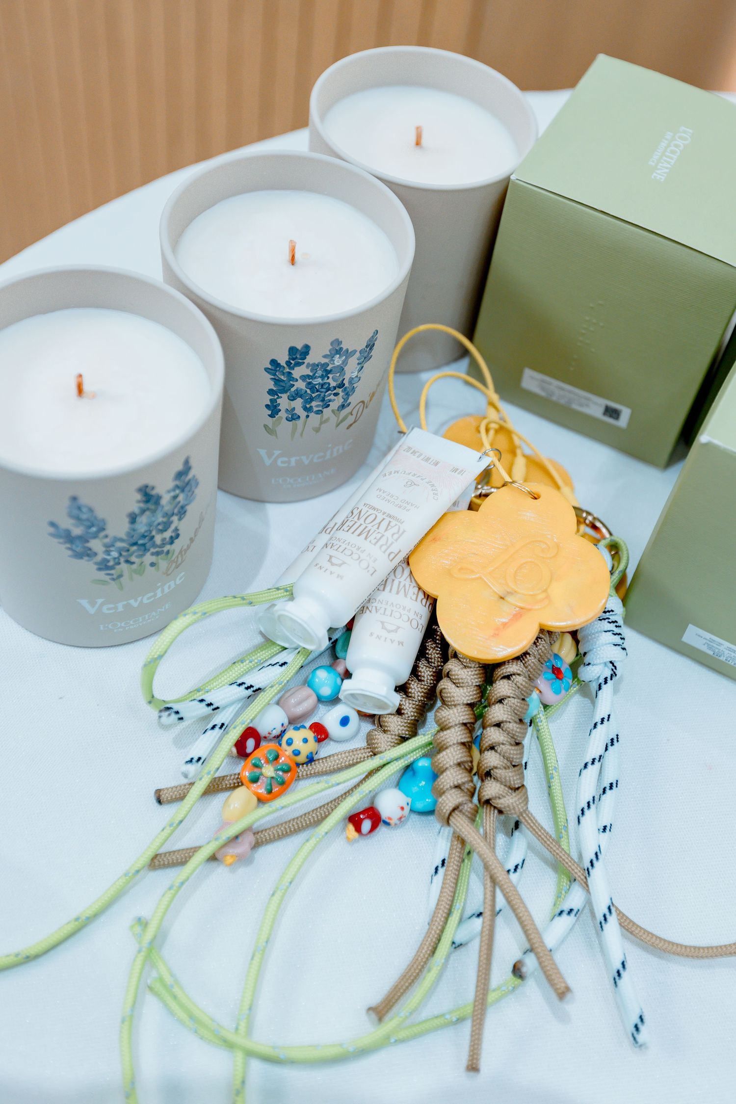 Scented candles with blue floral designs, decorative beaded charms, hand cream, and green gift boxes on white surface.