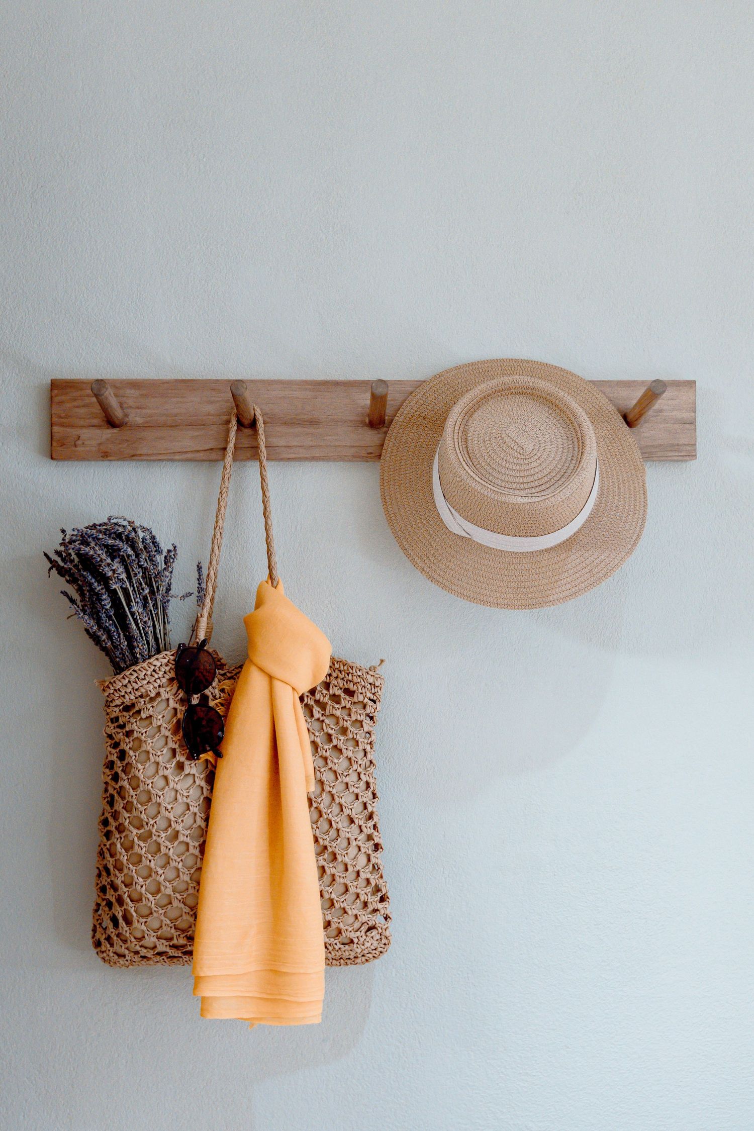 Wooden wall hook rack with woven bag, orange towel, lavender bouquet, sunglasses, and straw hat on light wall.