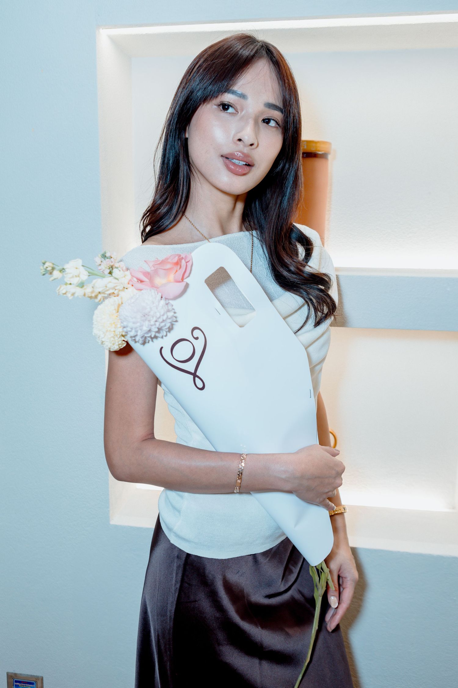 Young woman in white top and dark satin skirt holding a white floral gift bag with pink roses indoors.