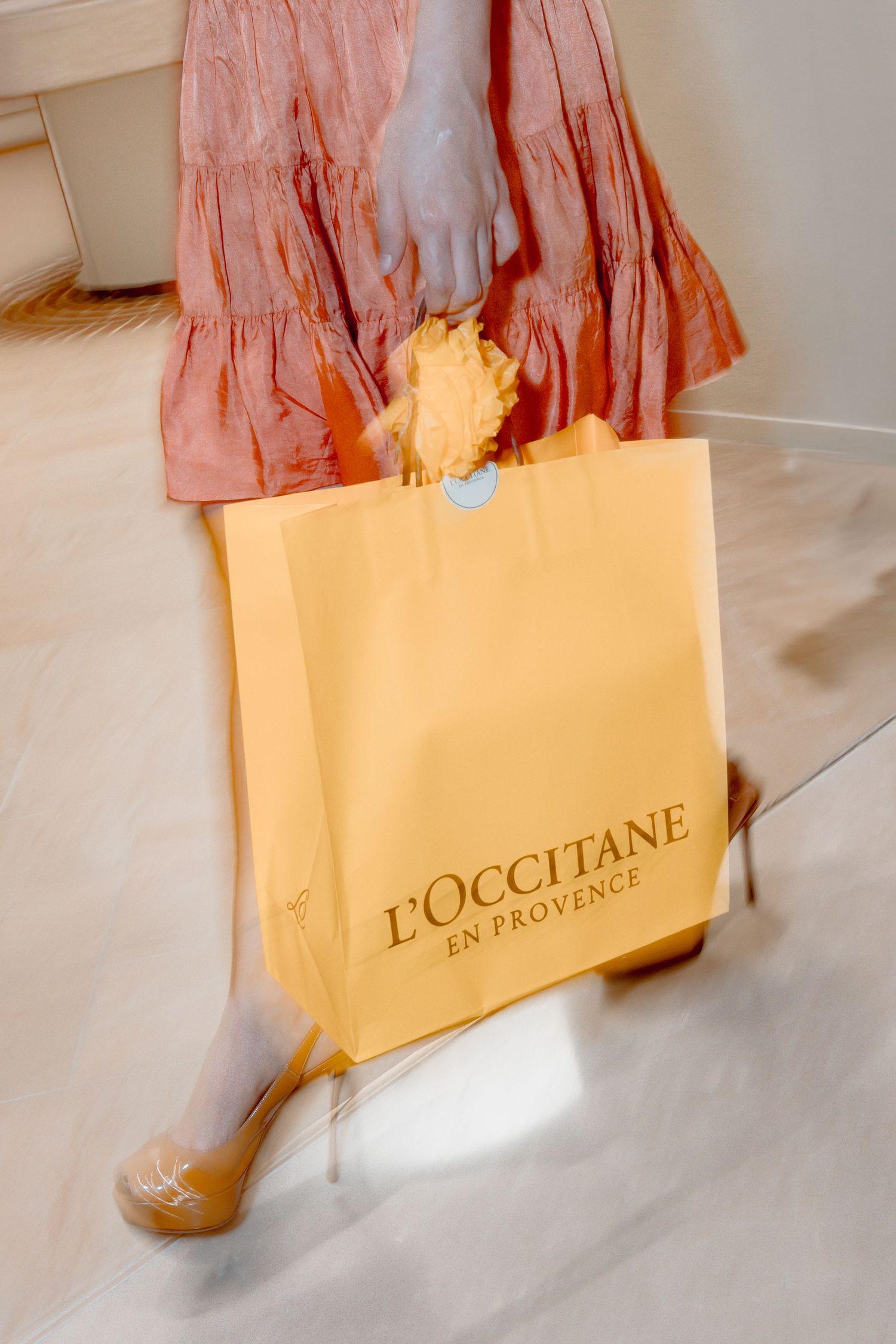 Woman in pink dress carrying a yellow L'Occitane en Provence shopping bag with a floral charm.