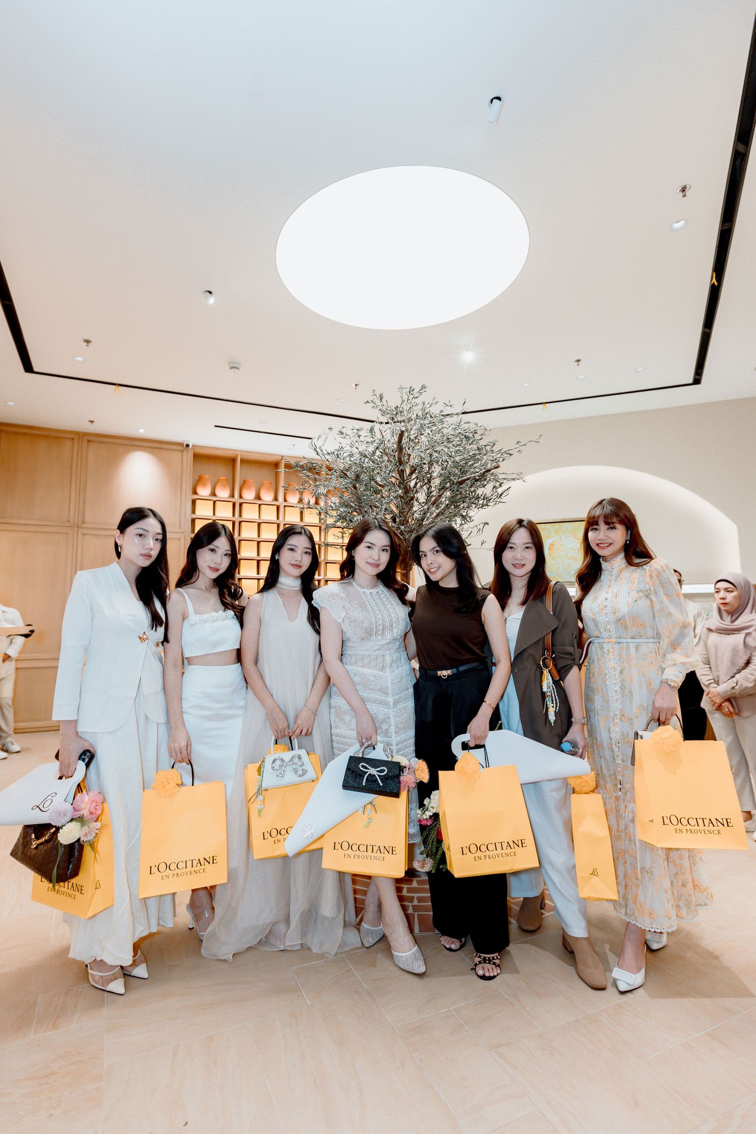 Group of stylish women posing together holding L'Occitane shopping bags in an elegant retail store interior.