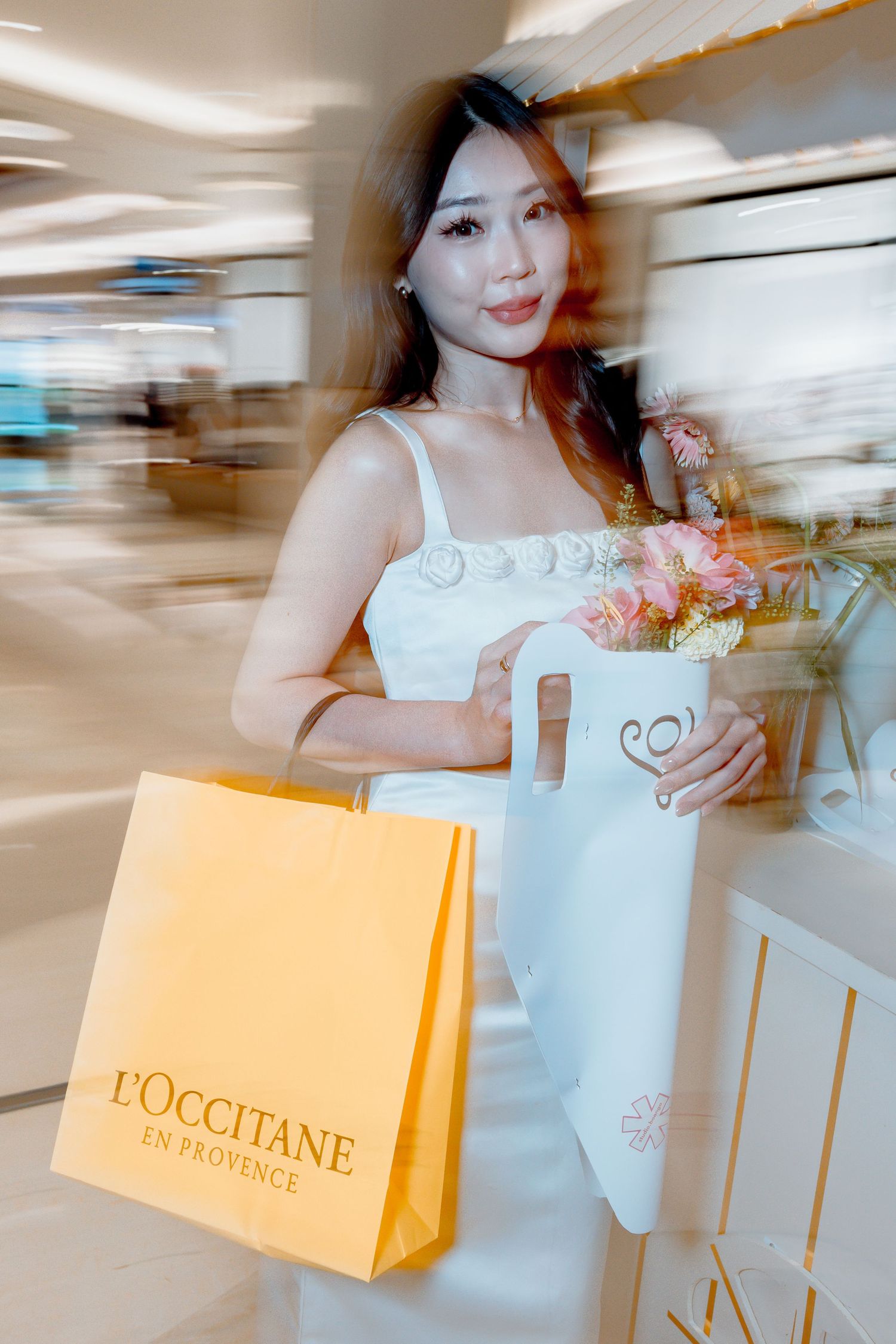 Woman in white dress holding L'Occitane shopping bag and floral vase in a luxury mall setting.