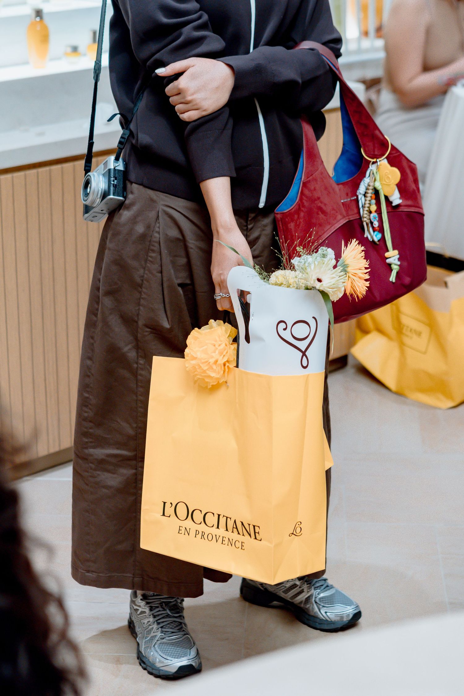 Person holding L'Occitane en Provence shopping bag with flowers, carrying red handbag and small camera.