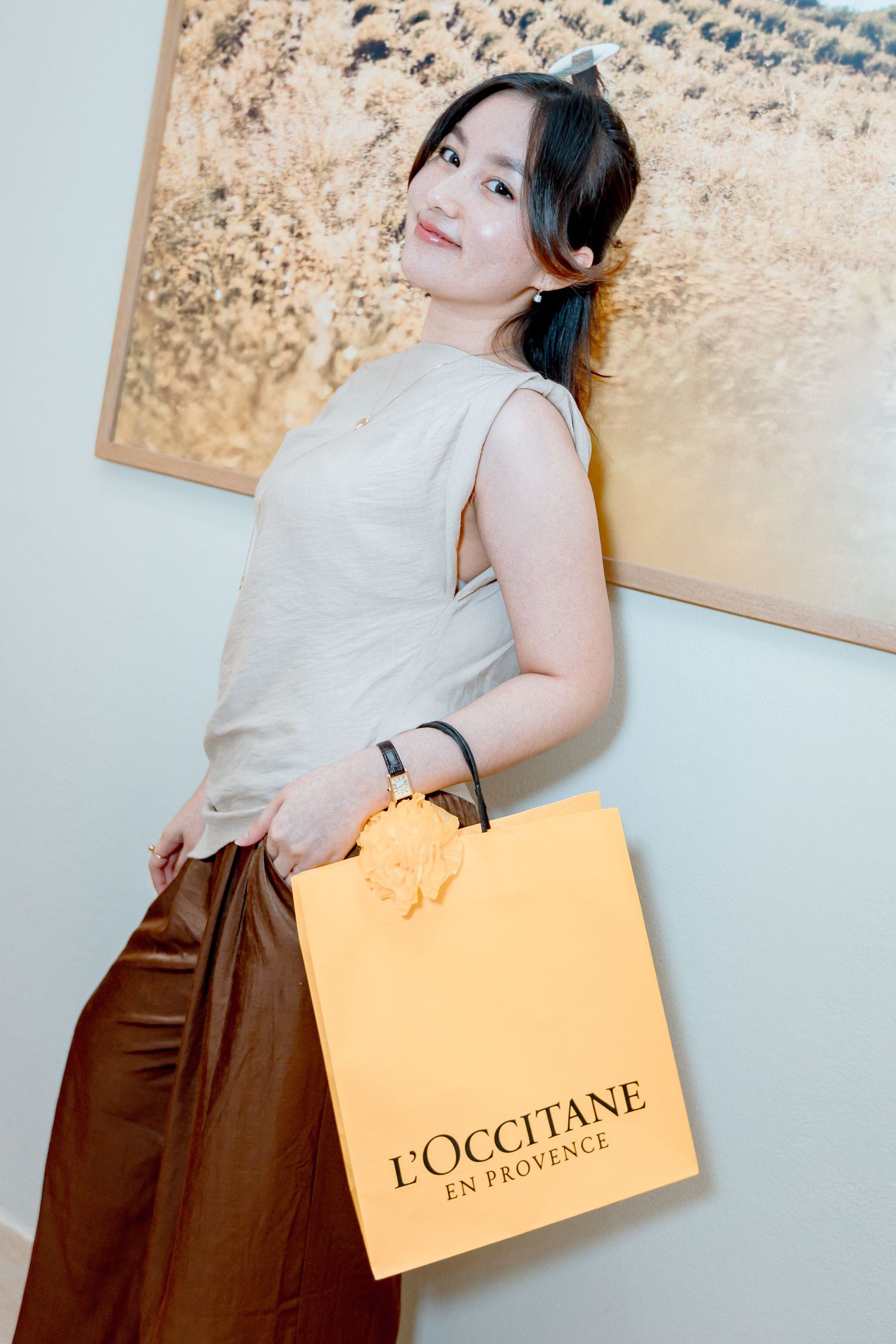 Young woman smiling and holding an L'Occitane en Provence orange shopping bag, wearing a beige top and brown satin pants.