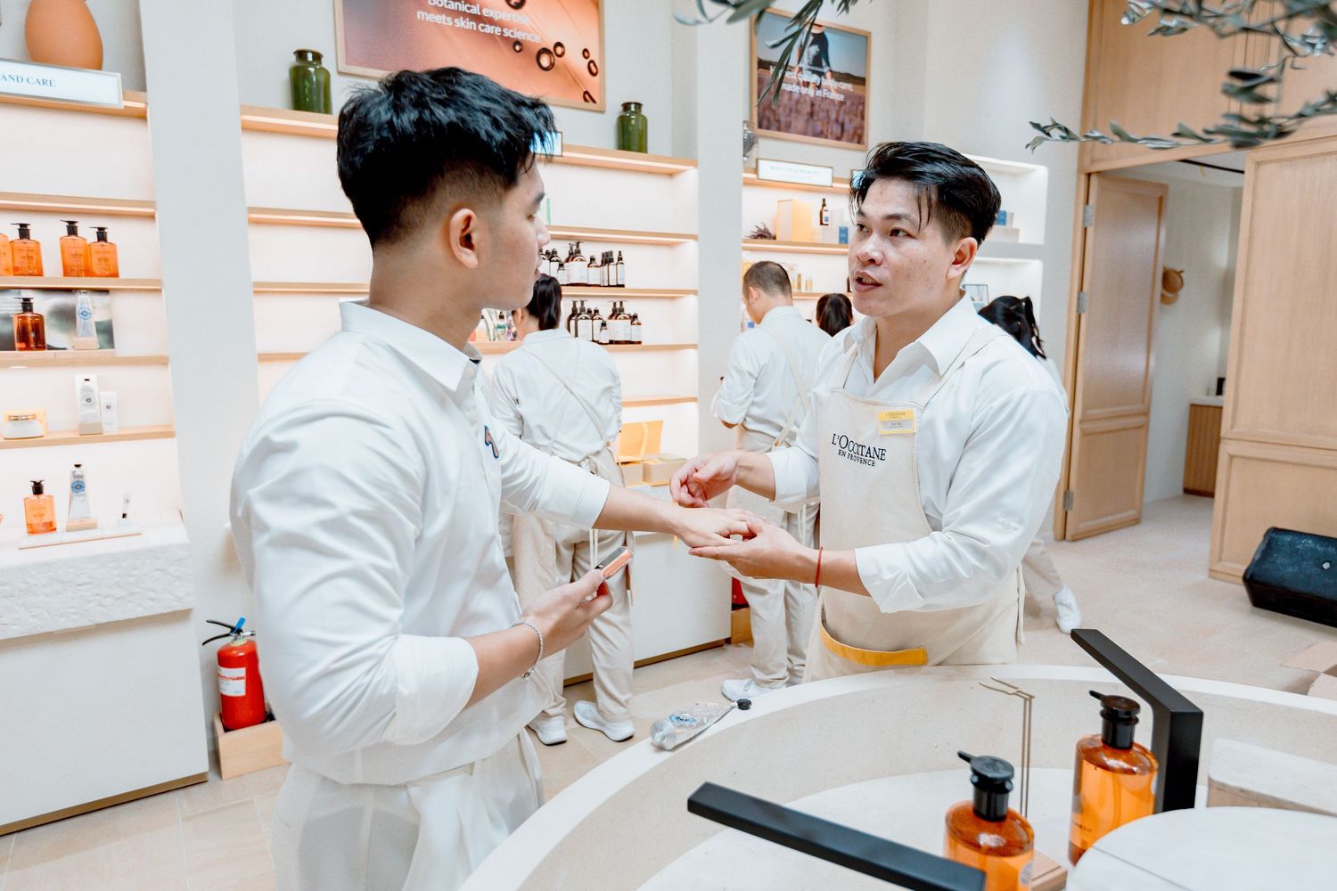 Two male spa staff in white uniforms discuss skincare products at a bright retail counter with shelves of beauty items.