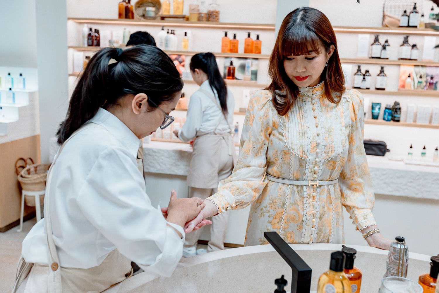 Two women interact at a perfume boutique counter, with fragrance bottles displayed on shelves in the background.