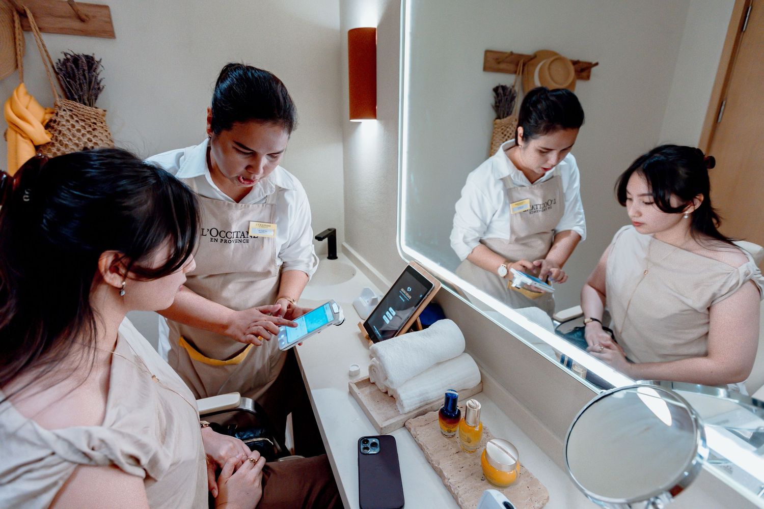 Female spa staff demonstrate skincare products to a client at a vanity mirror in a modern beauty salon.