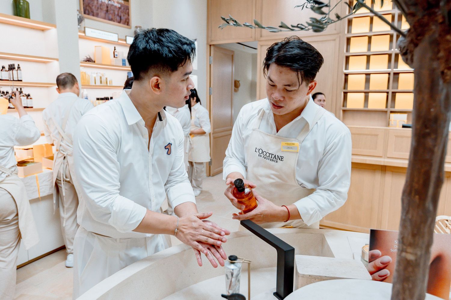 Two men in white shirts explore skincare products at an elegant L'Occitane beauty store counter.