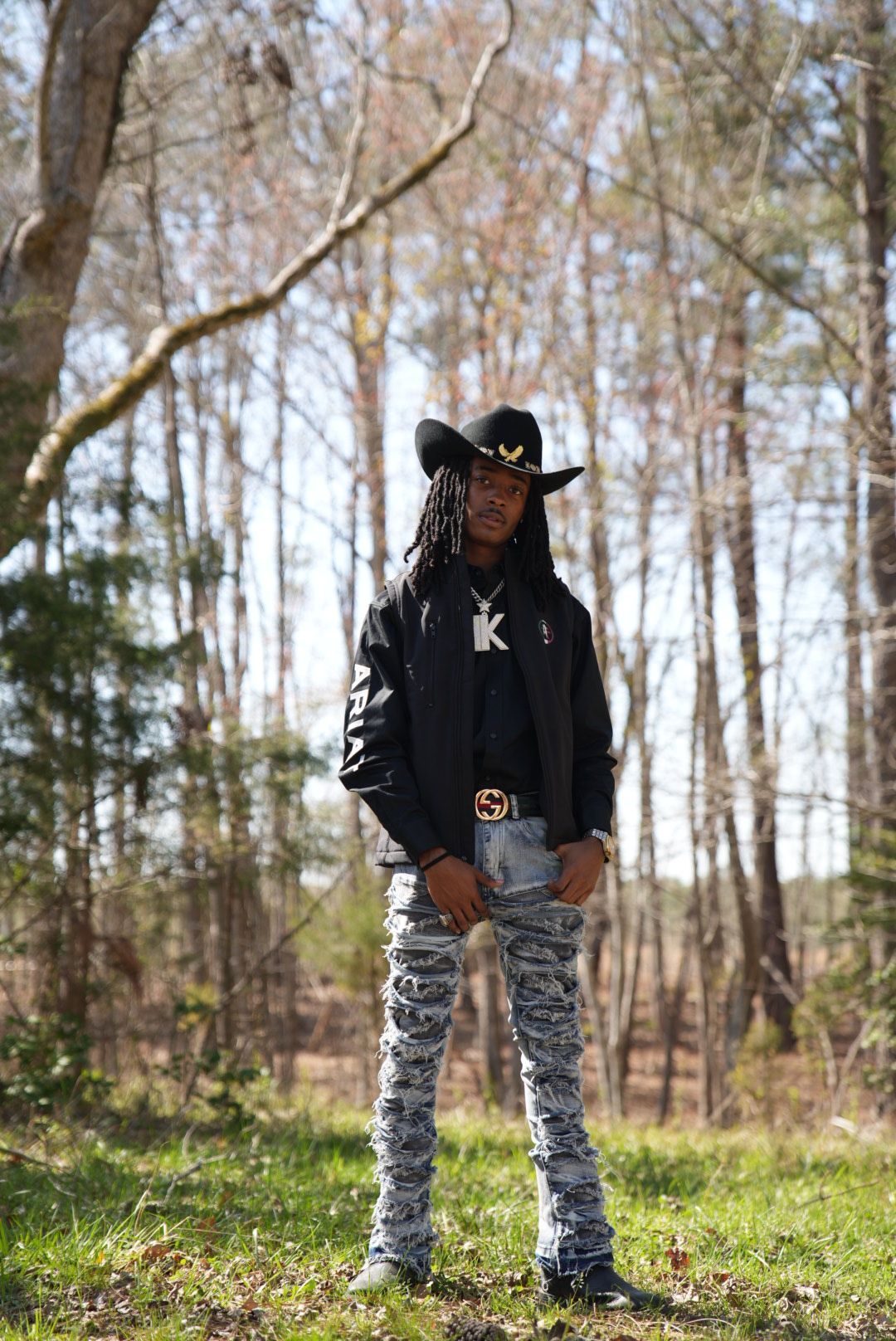 A young man in a black cowboy hat, jacket, and distressed stacked jeans stands on grass among tall trees on a sunny day.