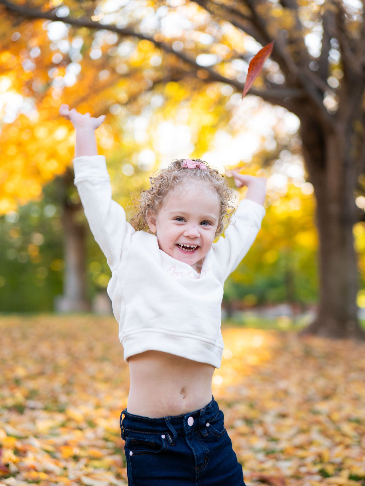 Happy young girl with curly hair raises arms joyfully among golden autumn leaves in a park.