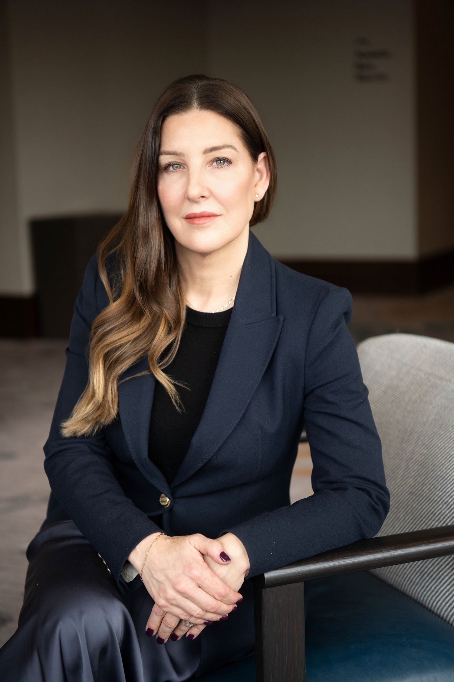 Professional portrait series of someone in a navy blazer and black top seated on a gray couch in an office setting.
