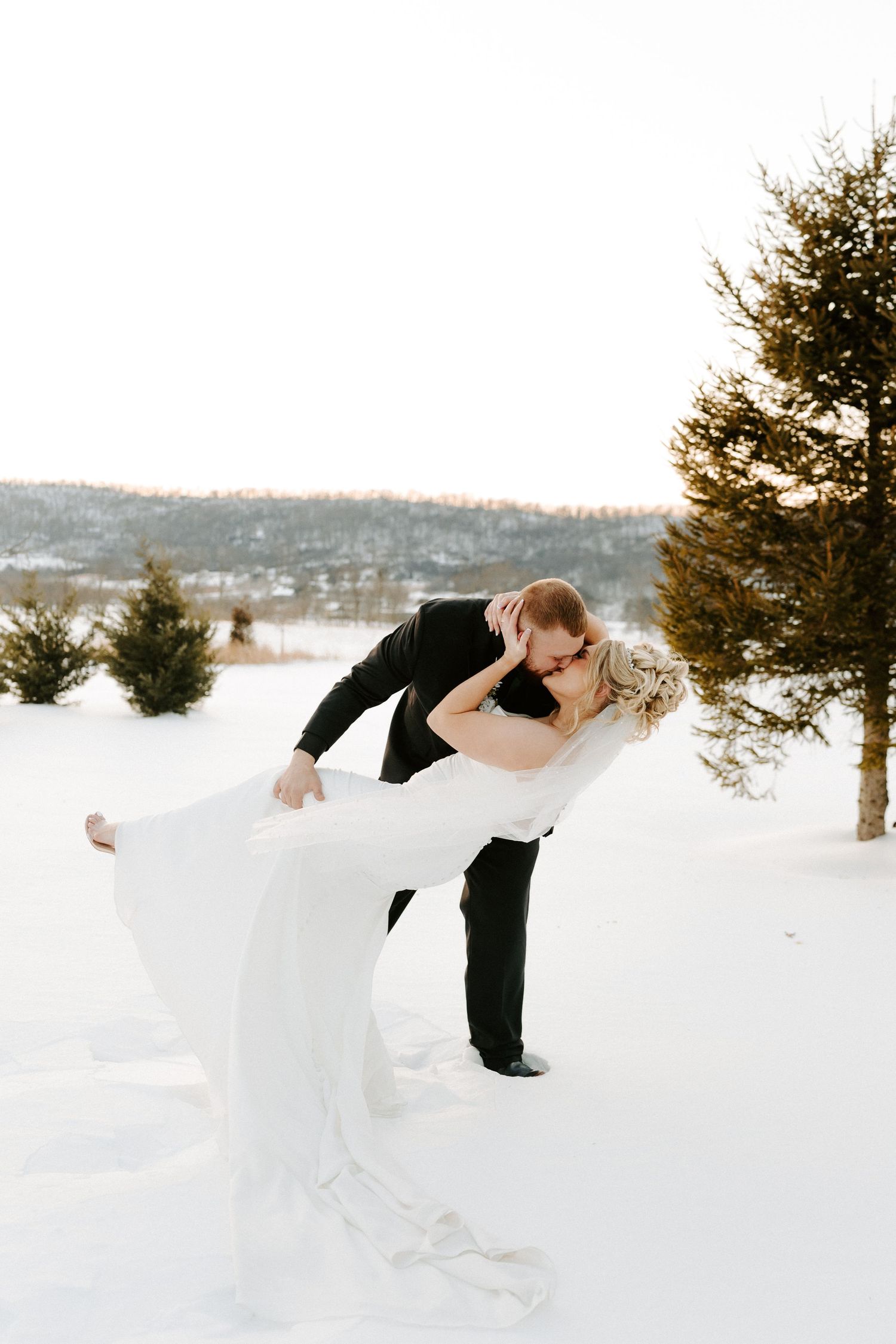 Groom dips bride for a kiss in snowy landscape with evergreen trees during winter wedding photos.