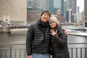 A couple in winter coats embraces while standing along a waterfront with city buildings in the background.