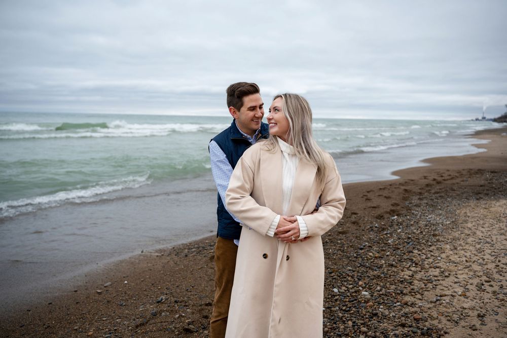 A couple shares romantic moments along a misty beach shoreline in neutral-toned outfits.