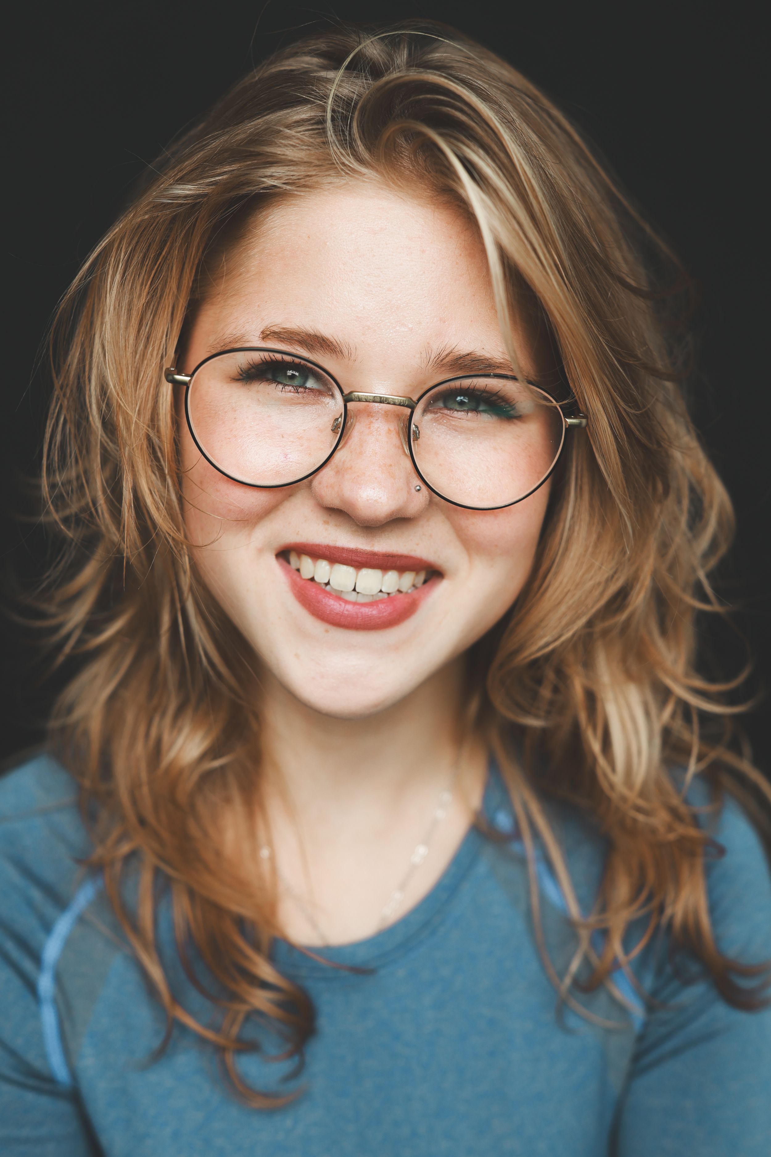 Smiling young woman with wavy red hair and round glasses wearing a blue shirt against a dark background.