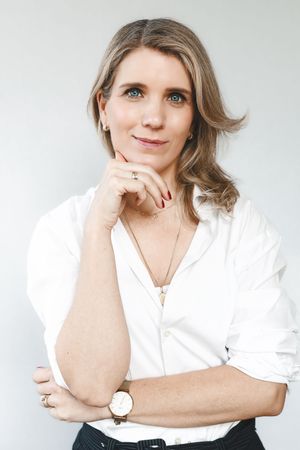 Professional portrait of a blonde woman in a white shirt posing with hand on chin against light background.