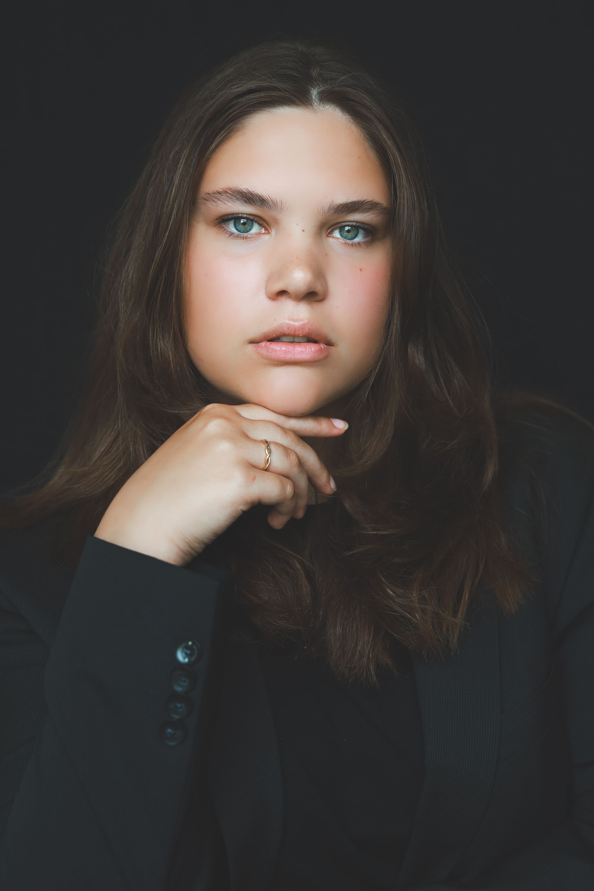 Portrait of a teenage girl with long brown hair and blue-green eyes in a black jacket against dark background.