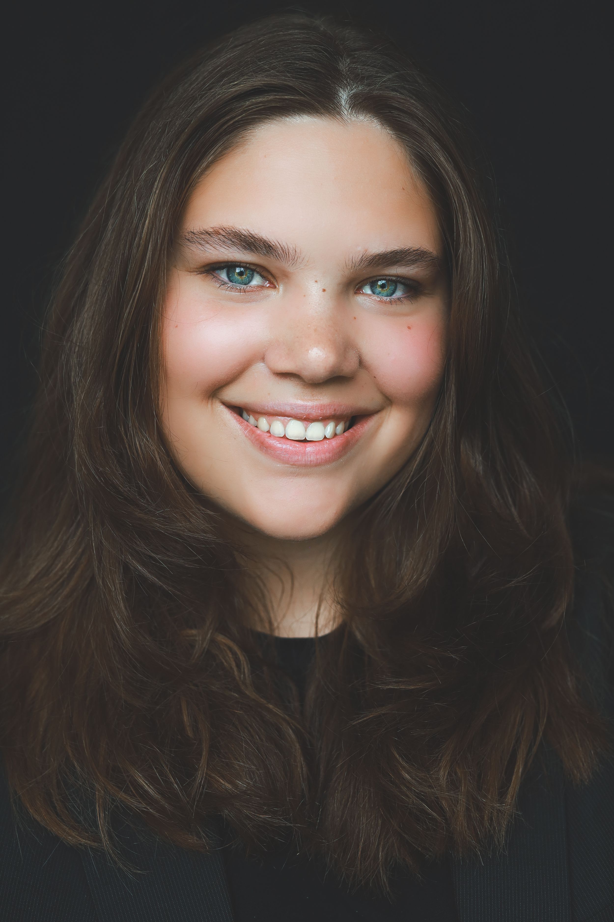 Smiling teenage girl with long brown hair and blue eyes wearing a dark top against a black background.