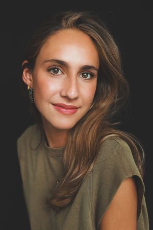 Young woman with brown hair and green eyes wearing a green top and gold earrings against dark background.
