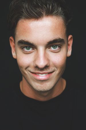 Headshot of a smiling young man with green eyes and short dark hair against a black background.
