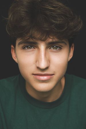 Portrait of a teenage boy with curly brown hair and hazel eyes wearing a green shirt on dark background.