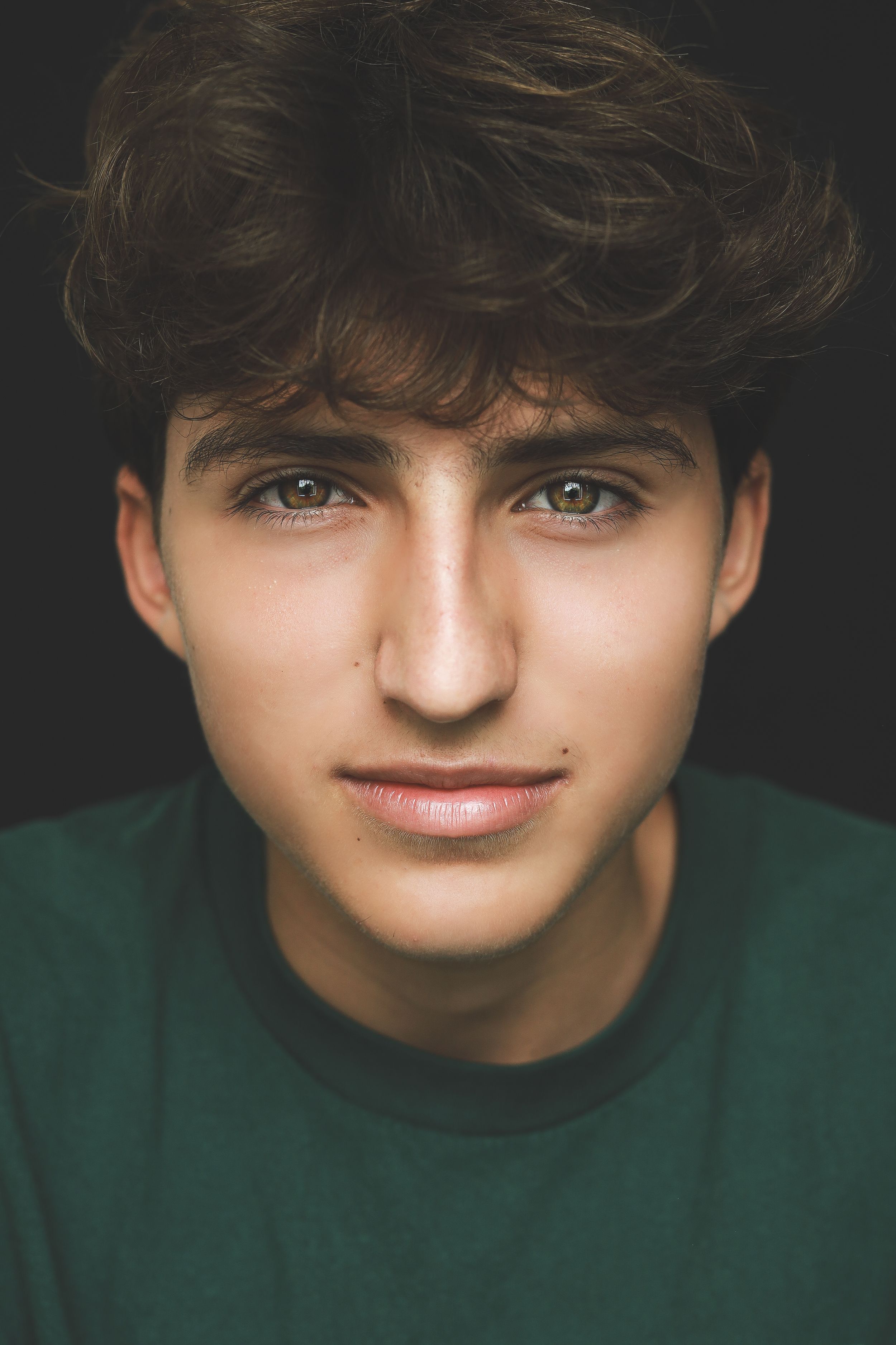 Portrait of a teenage boy with curly brown hair and hazel eyes wearing a green shirt on dark background.