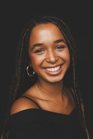 Joyful young woman with braided hair and hoop earring smiling brightly in a black off-shoulder top.