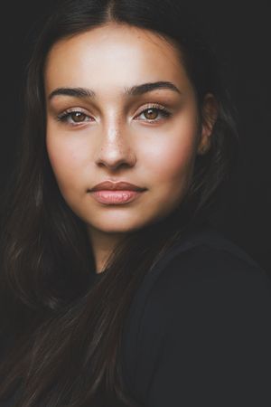 Close-up portrait of a young brunette woman with freckles and brown eyes against a dark background.